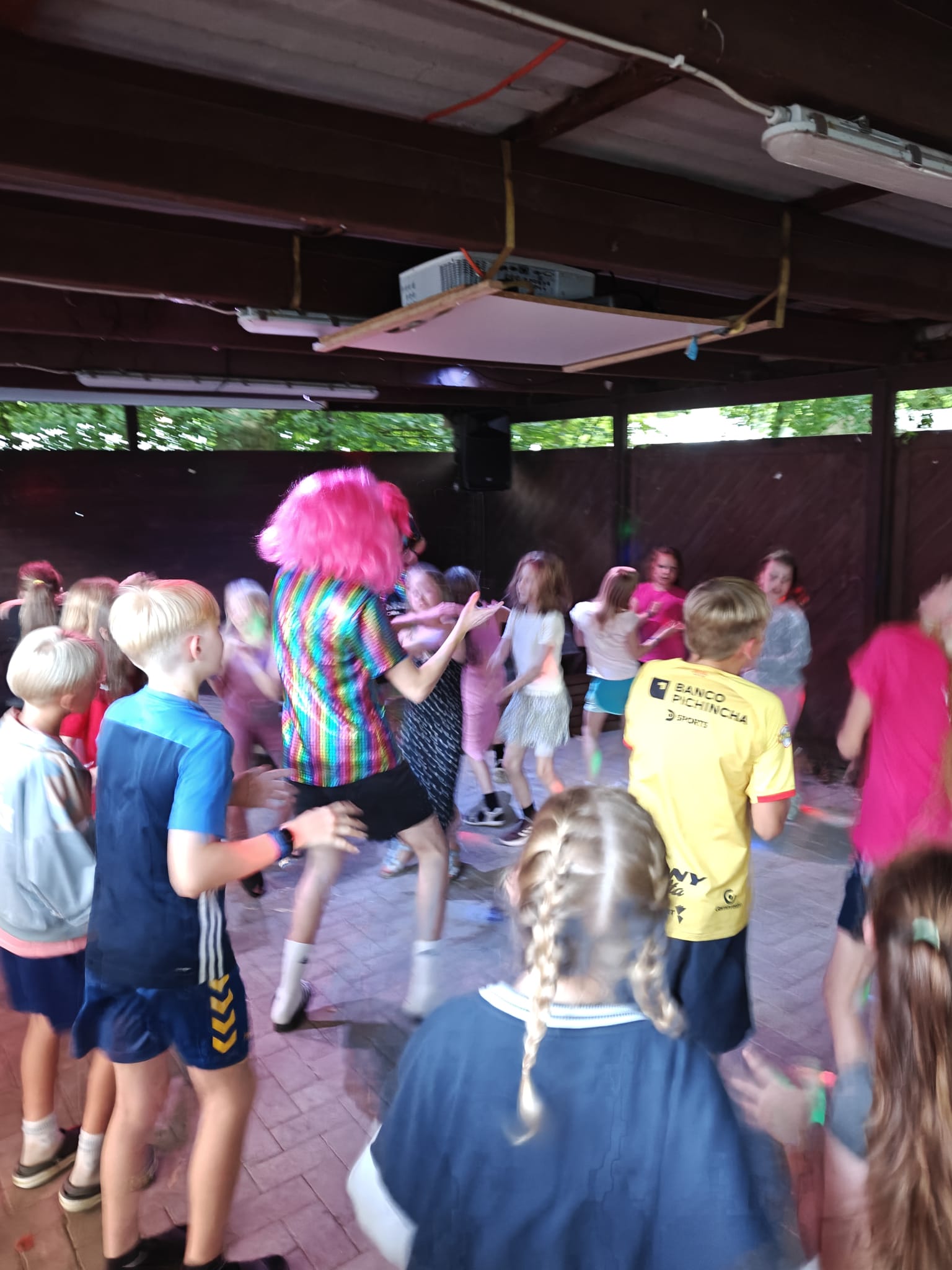 Zeltlager Meesdorf 2026 - Tag 2 - Disco Kinder tanzen in einem überdachten Bereich mit einer Person in einem bunten Hemd und pinker Perücke
