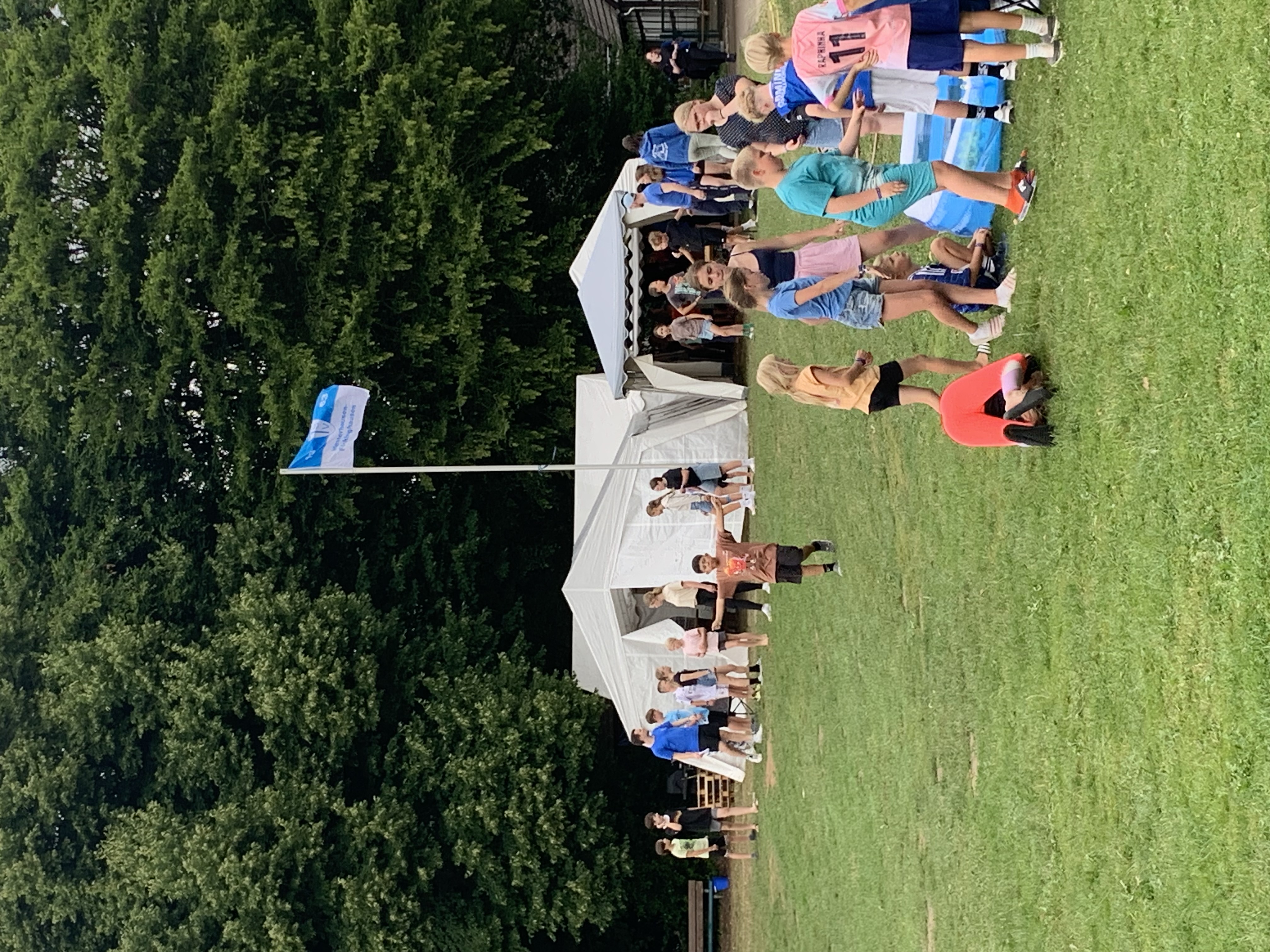 Zeltlager Meesdorf 2026 - Tag 2 - Zeltplatz mit Fahne Kinder spielen auf einer Wiese vor zwei Zelten, im Hintergrund Bäume und eine Fahne auf einem Mast