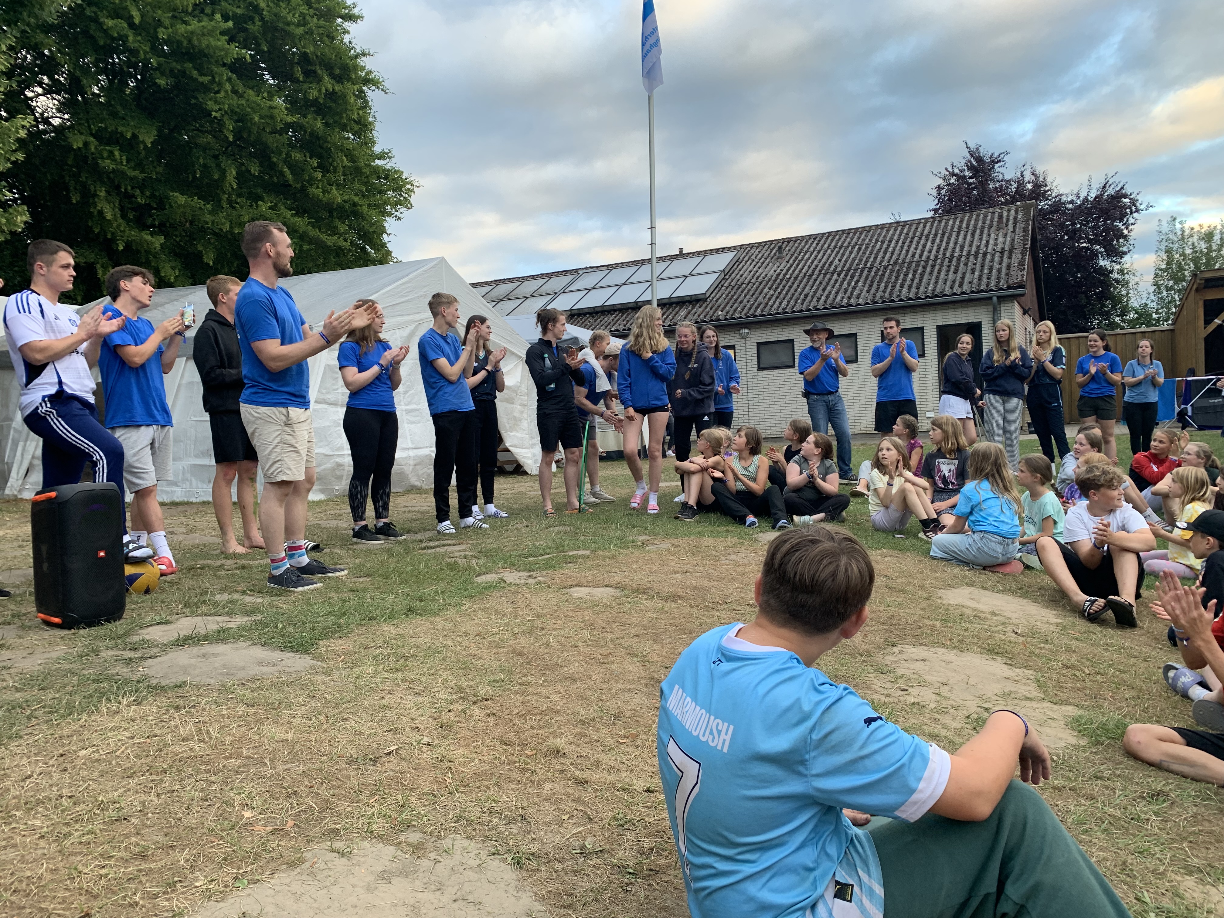 Zeltlager Meesdorf 2026 - Tag 4 - Runde Gruppe von Jugendlichen und Erwachsenen steht und sitzt auf einer Wiese vor einem Gebäude mit Solarpaneelen auf dem Dach, einige klatschen, im Vordergrund sitzt eine Person mit blauem T-Shirt mit der Aufschrift 'Westerhausen'
