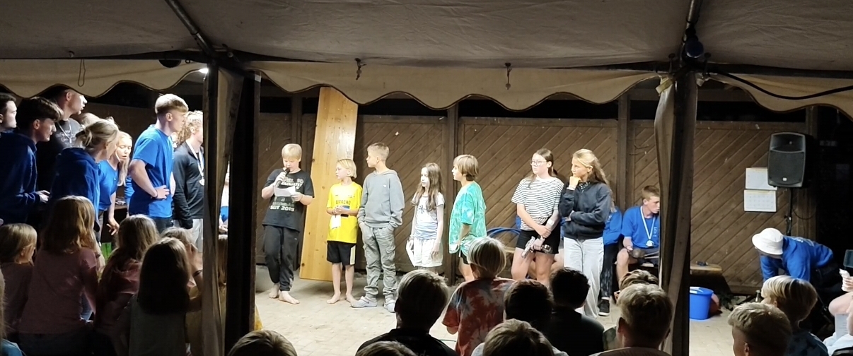 Zeltlager Meesdorf 2026 - Tag 5 - 1 Gruppe von Kindern und Jugendlichen steht auf einer Bühne unter einem Zelt, davor sitzt Publikum im Dunkeln