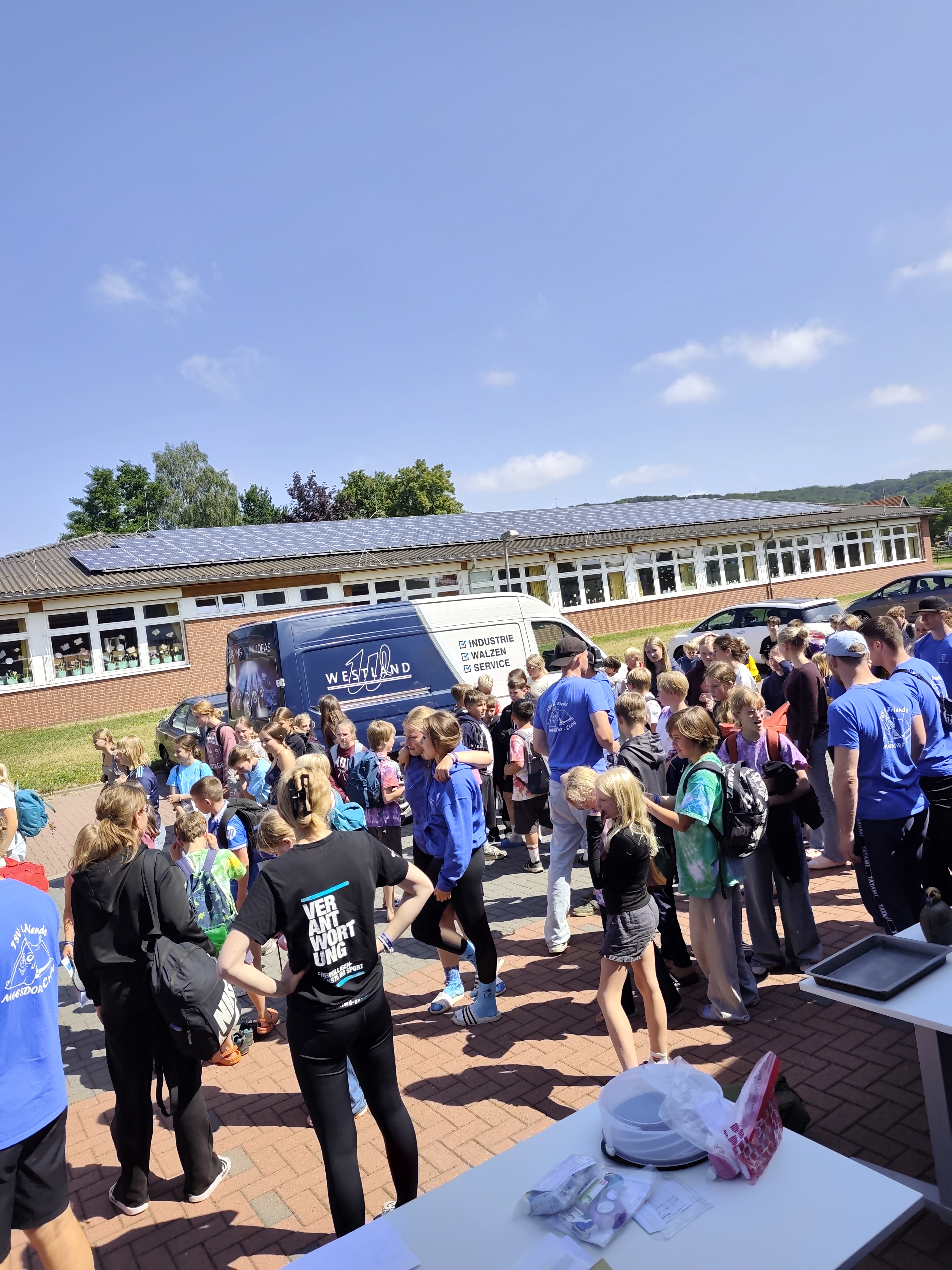 Zeltlager Meesdorf - Tag 6 Gruppe von Kindern und Jugendlichen in blauen T-Shirts versammelt auf einem Schulhof vor einem Gebäude mit Solarpaneelen auf dem Dach