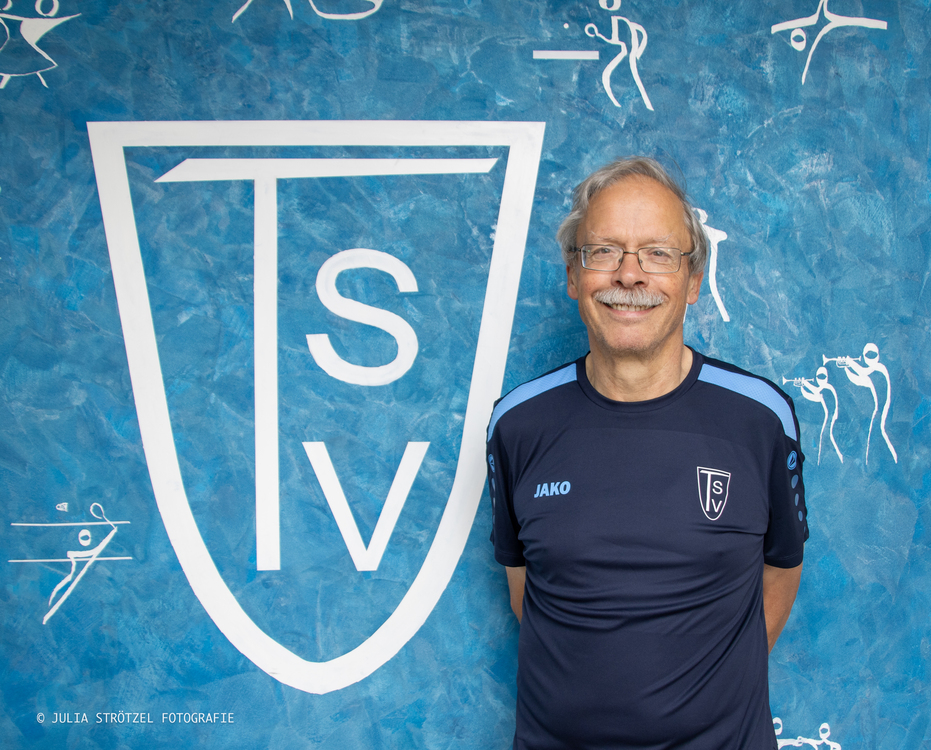 Mann in dunkelblauem Sportshirt mit TSv-Logo steht vor blauem Hintergrund mit großem weißem TSV-Wappen und kleinen weißen Sportler-Silhouetten