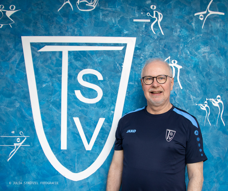 Ingo Timmermeister Mann in dunkelblauem Sportshirt mit JAKO-Logo und TSV-Wappen vor blauem Hintergrund mit großem weißem TSV-Wappen und weißen Sportpiktogrammen.