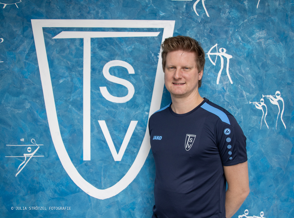 Jan Plegge Mann in dunkelblauem Sportshirt mit TSV-Logo steht vor blauem Hintergrund mit großem weißem TSV-Wappen und weißen Sportpiktogrammen.