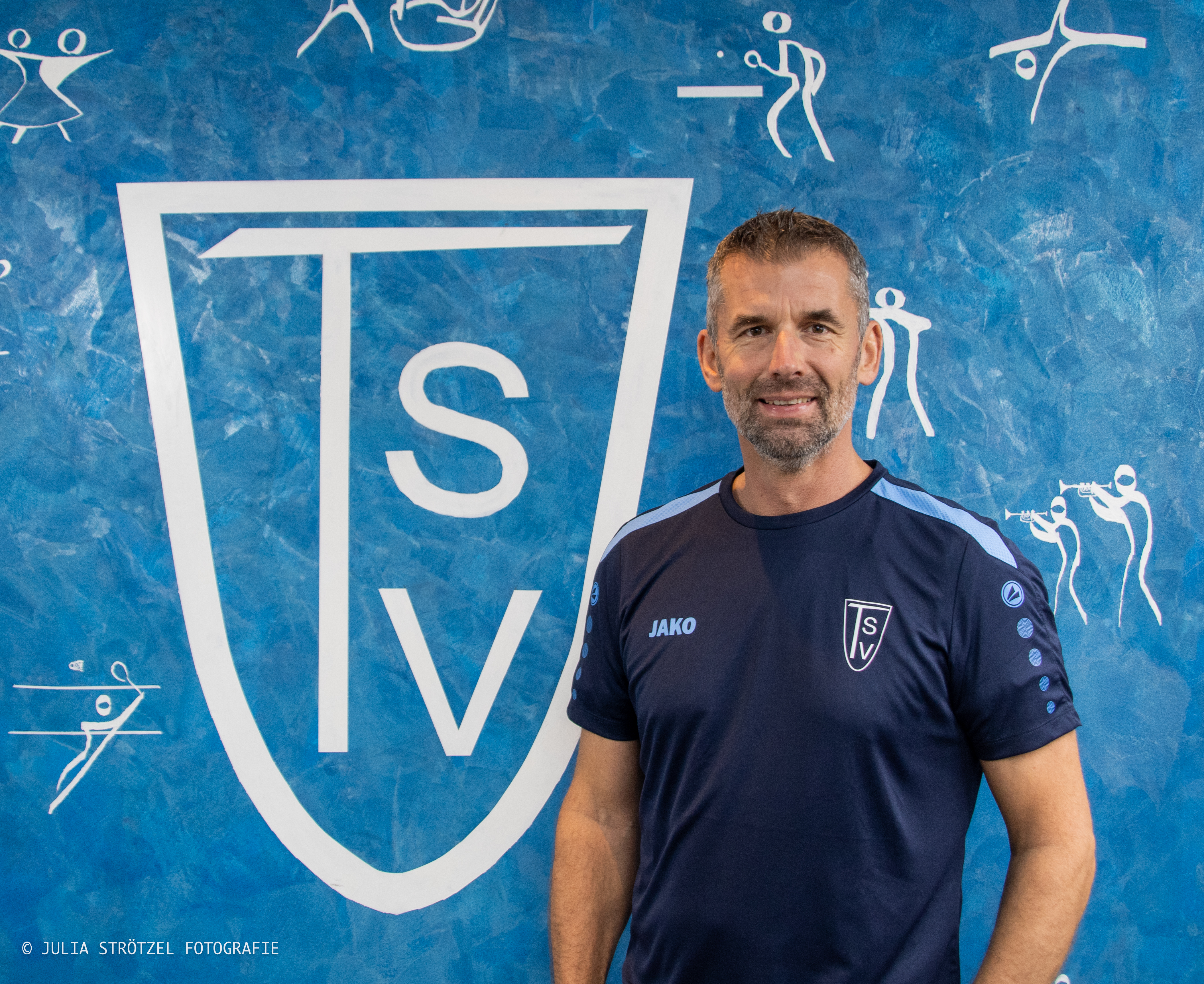 Mann in blauem TSV-Sportshirt steht vor blauem Hintergrund mit großem weißem TSV-Logo und weißen Sportpiktogrammen.
