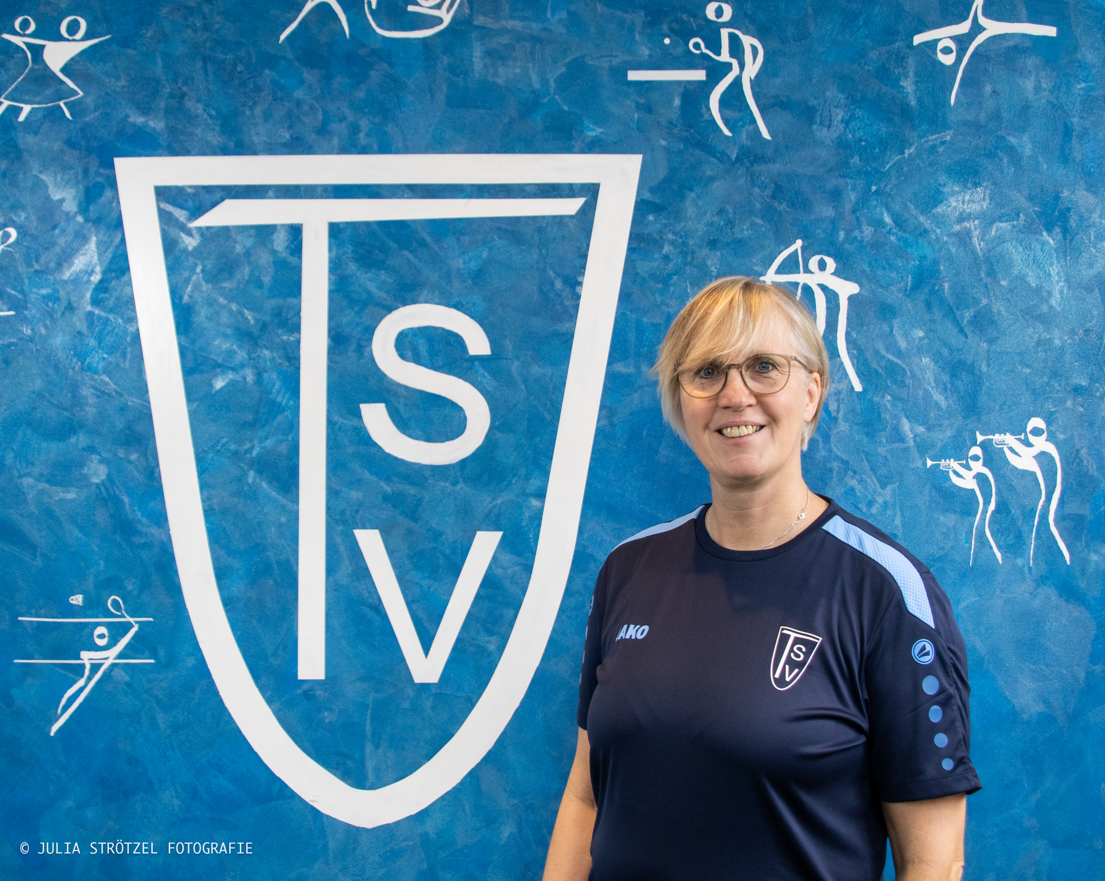 Person in dunkelblauem Sportshirt mit hellblauen Ärmeln steht vor blauem Hintergrund mit großem weißem TSV-Logo und weißen Sportpiktogrammen.