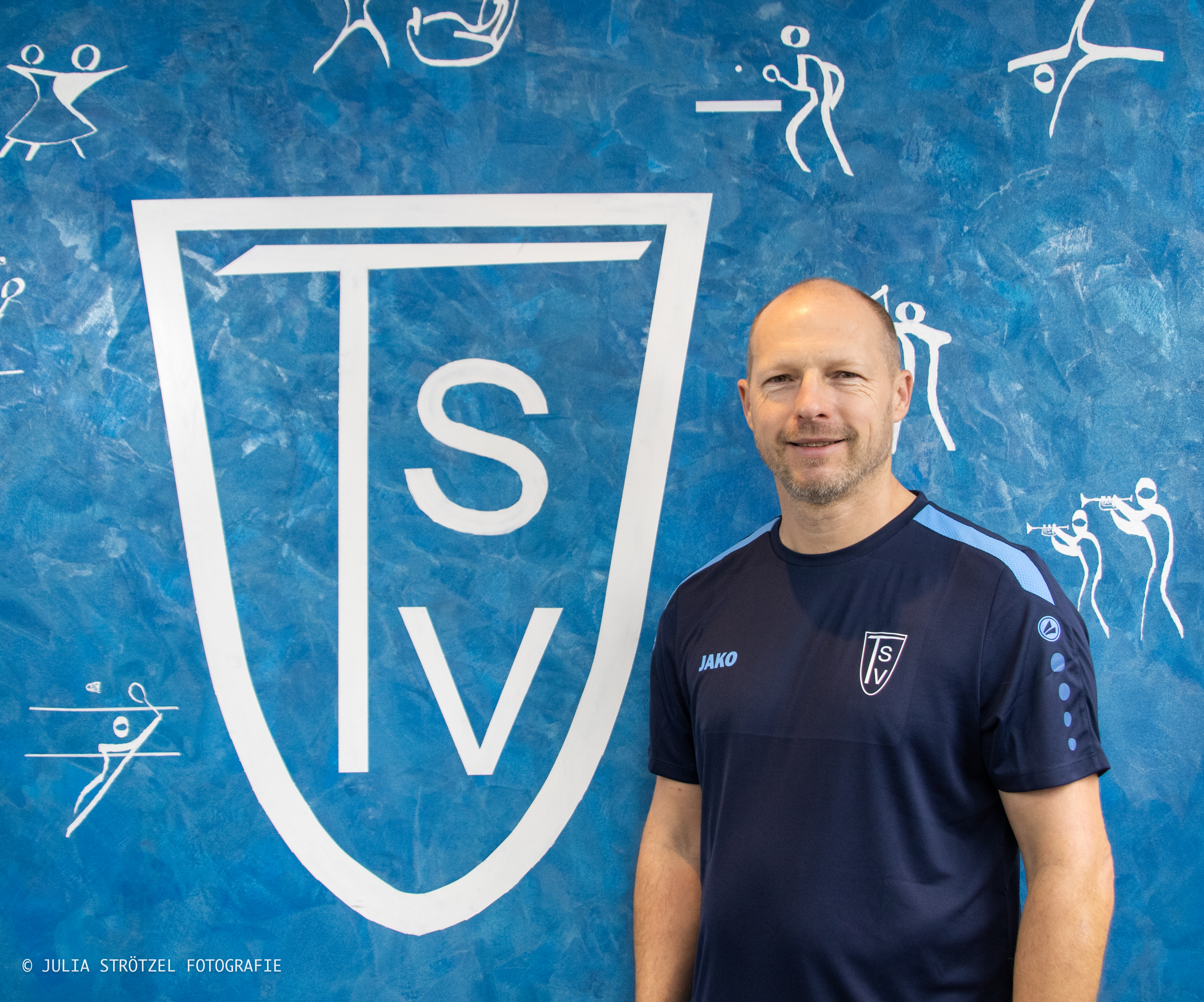 Mann in dunkelblauem TSV-Sportshirt steht vor blauem Hintergrund mit großem weißem TSV-Logo und weißen Sportpiktogrammen.