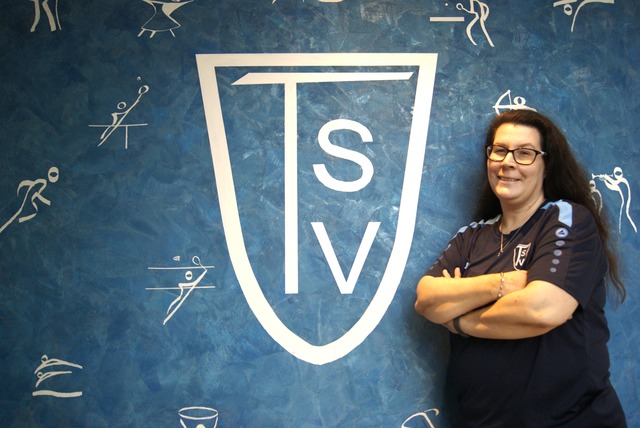Person weiblich,  Michaela Strötzel steht mit verschränkten Armen vor einer blauen Wand mit großem TSV-Logo und kleinen Sportpiktogrammen
