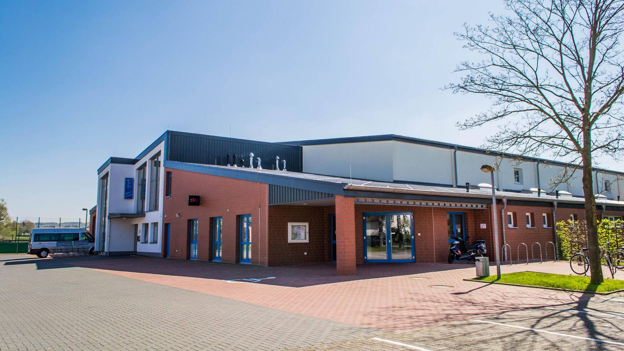 Sportzentrum am Ochsenweg Eingangsbereich eines modernen Gebäudes mit rotem Backstein, großen Fenstern und Flachdach, davor gepflasterter Platz und Baum ohne Blätter.