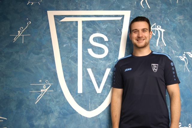 Person männlich, Nils auf der Masch steht mit verschränkten Armen vor einer blauen Wand mit großem TSV-Logo und kleinen Sportpiktogrammen